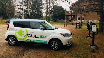 A white Kia Desrosiers EV charging at a public charging station in a grassy area with trees and a wooden structure in the background.