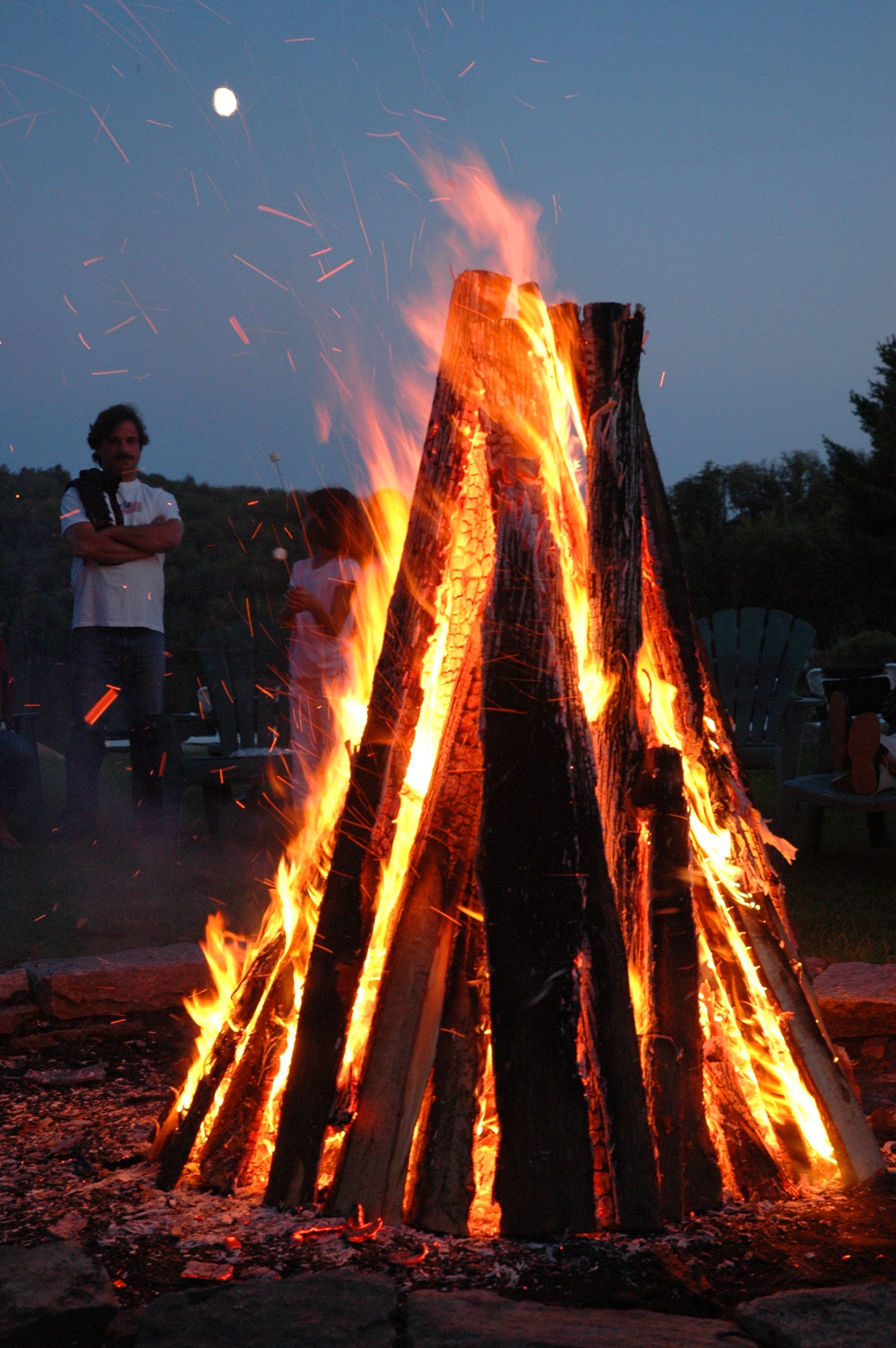 Ein Lagerfeuer mit vertikal gestapelten Holzscheiten, umringt von Personen und Stühlen.