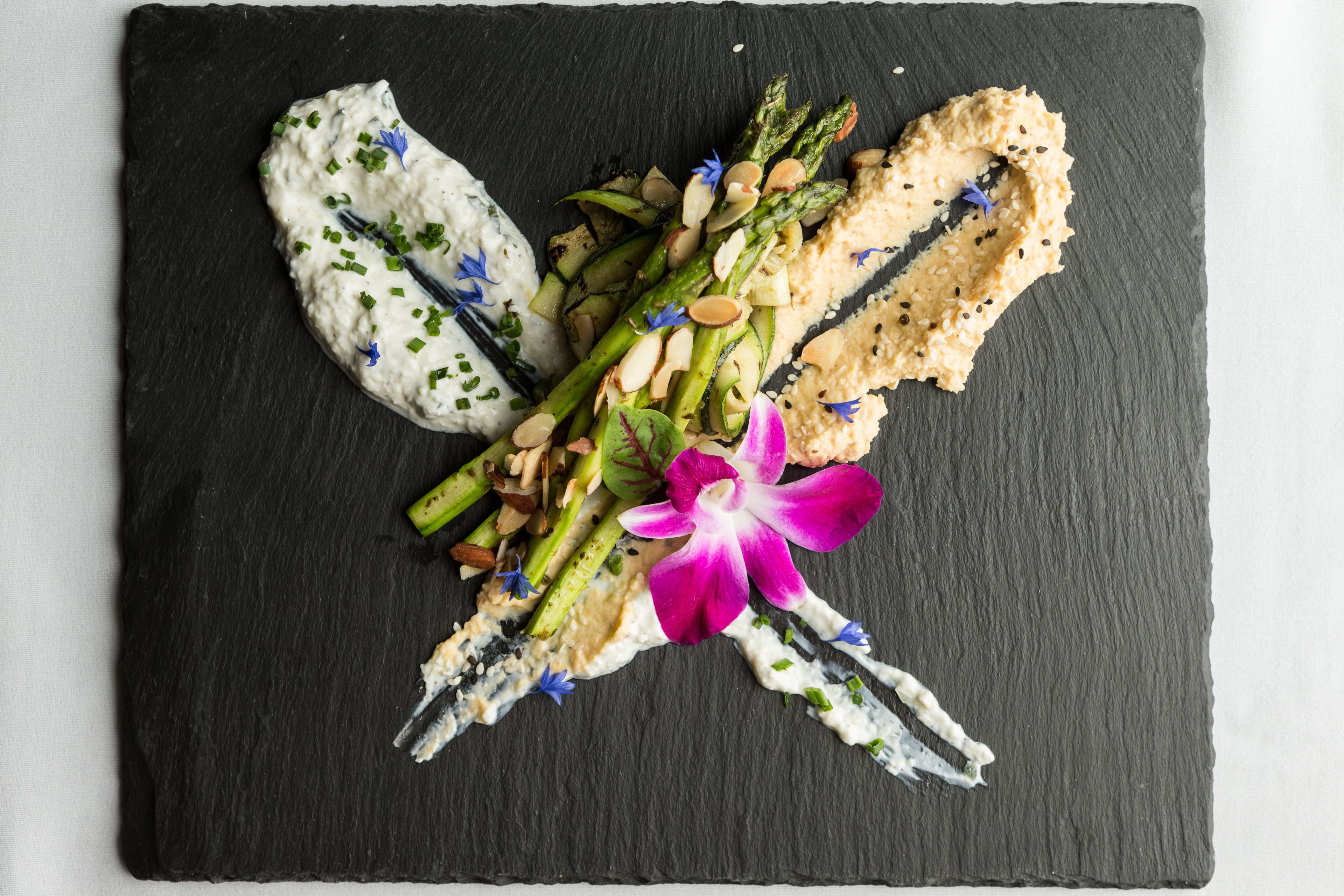 A black slate board with a food arrangement featuring asparagus, almonds, and a purple flower.