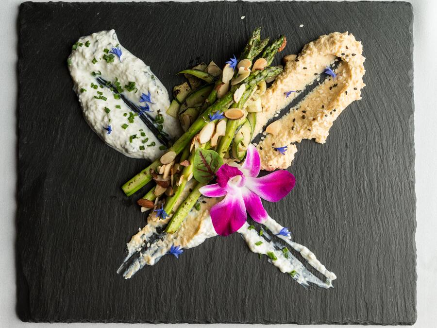 A black slate board with a food arrangement featuring asparagus, almonds, and a purple flower.