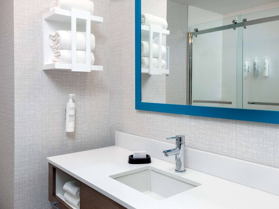 A modern bathroom with a white sink, chrome faucet, and a large mirror with a blue frame. There are shelves with towels and toiletries.