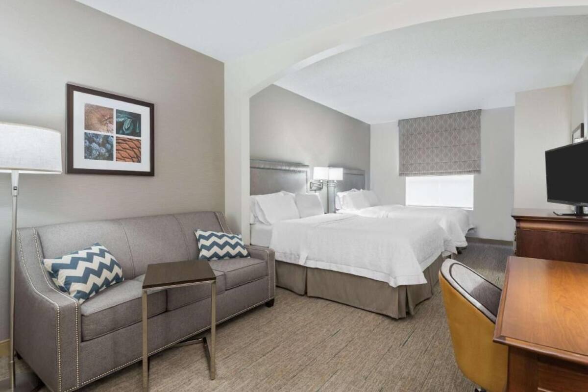 A hotel room with a sofa, two beds, a desk, a TV, and a framed picture on the wall.