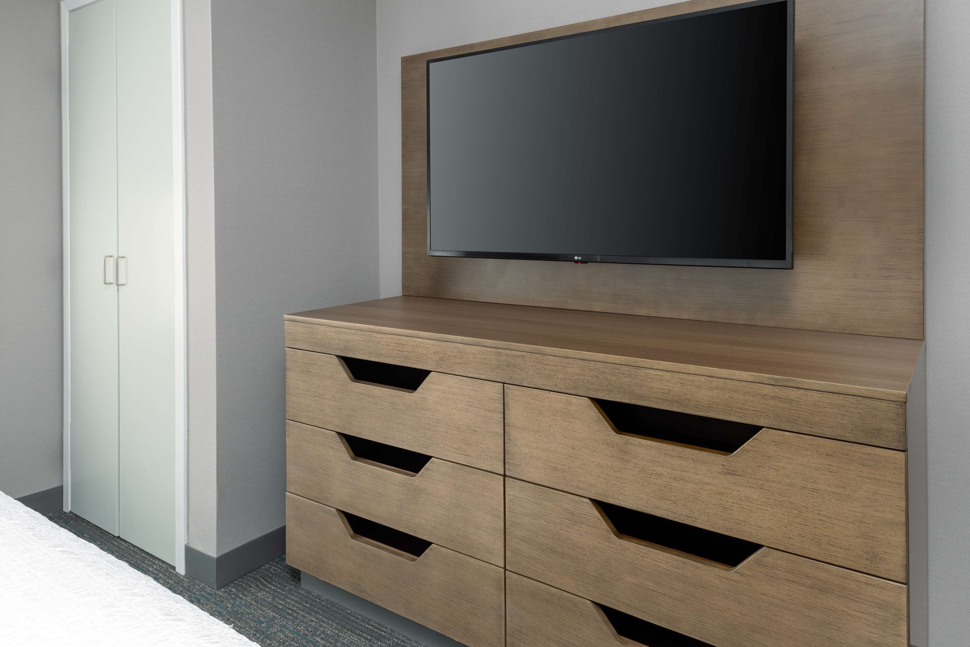 Een moderne kamer met een grote flatscreen-tv aan een houten kast, een garderobe met een spiegeldeur, en een vloerbedekking.