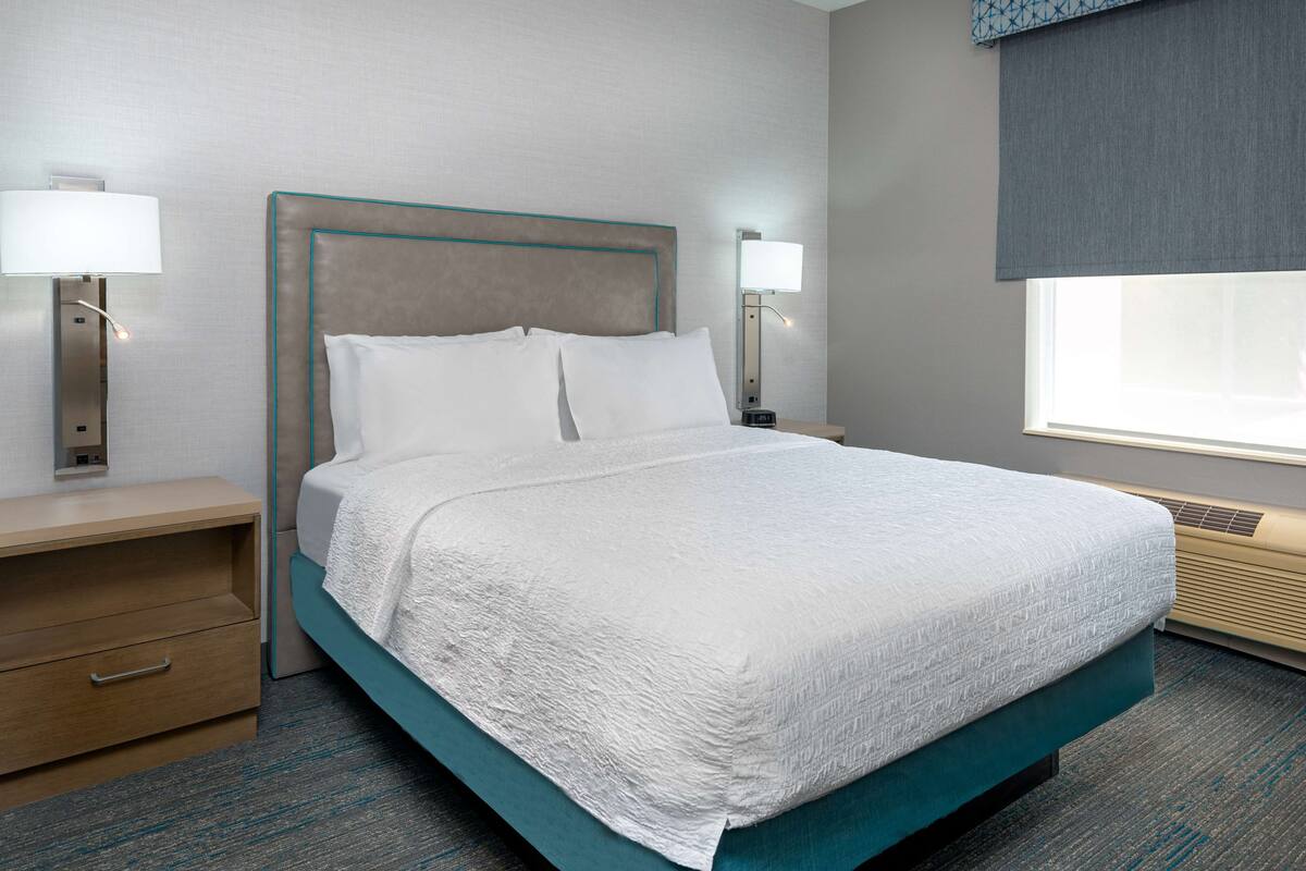 A neatly made bed with a teal frame and white bedding, a bedside table with a lamp, and a window with a view.