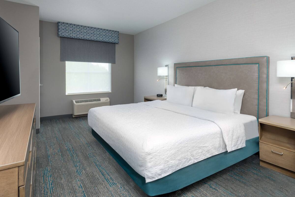A hotel room with a bed, bedside tables, a TV, a window with blinds, and a wall-mounted air conditioner.