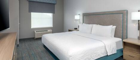A hotel room with a bed, bedside tables, a TV, a window with blinds, and a wall-mounted air conditioner.