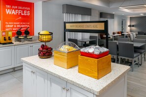 A breakfast buffet area with waffle makers, fruit bowls, and a sign indicating breakfast service. - Hampton Inn Boston Bedford Burlington (Billerica)