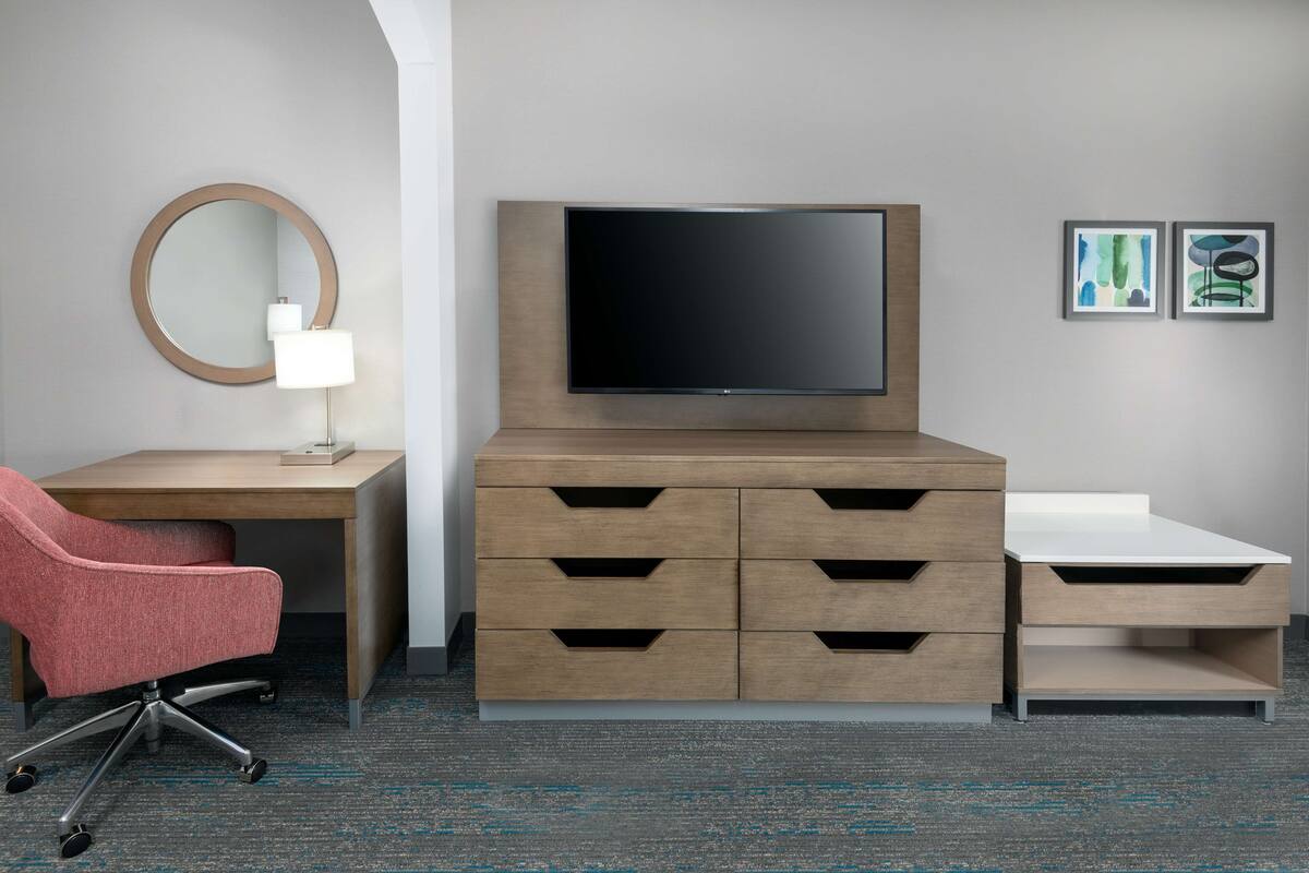 A hotel room with a wooden desk, a red office chair, a flat-screen TV, a bedside table, a mirror, and two framed artworks on the wall.