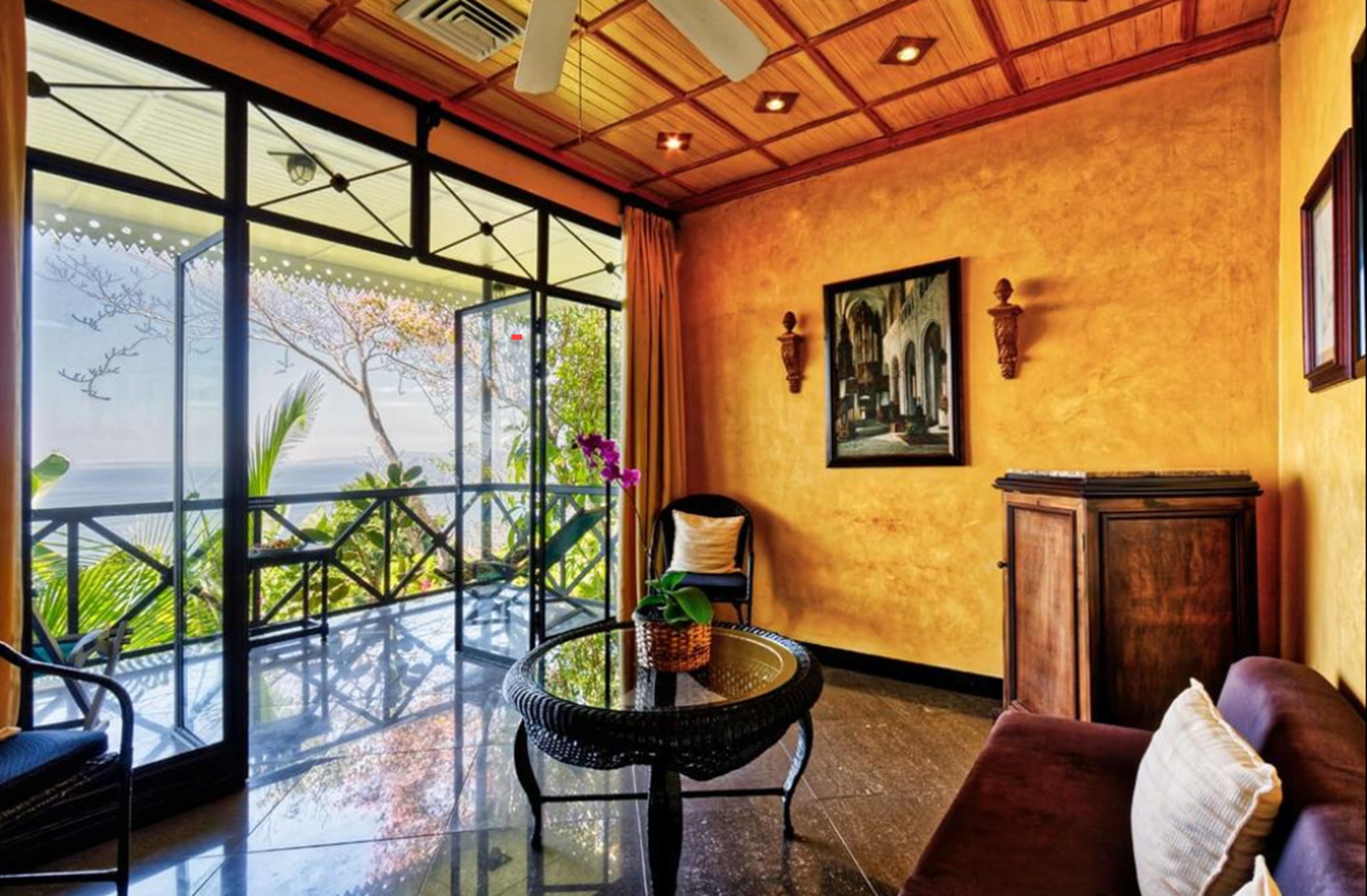 A room with a large glass door leading to a balcony with a view of palm trees. There is a round table with a vase of flowers, a cushioned chair, and a wooden cabinet.