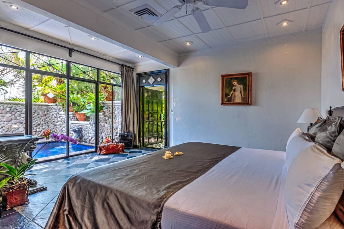 A bedroom with a large bed, a painting on the wall, and a view of a garden through the window.