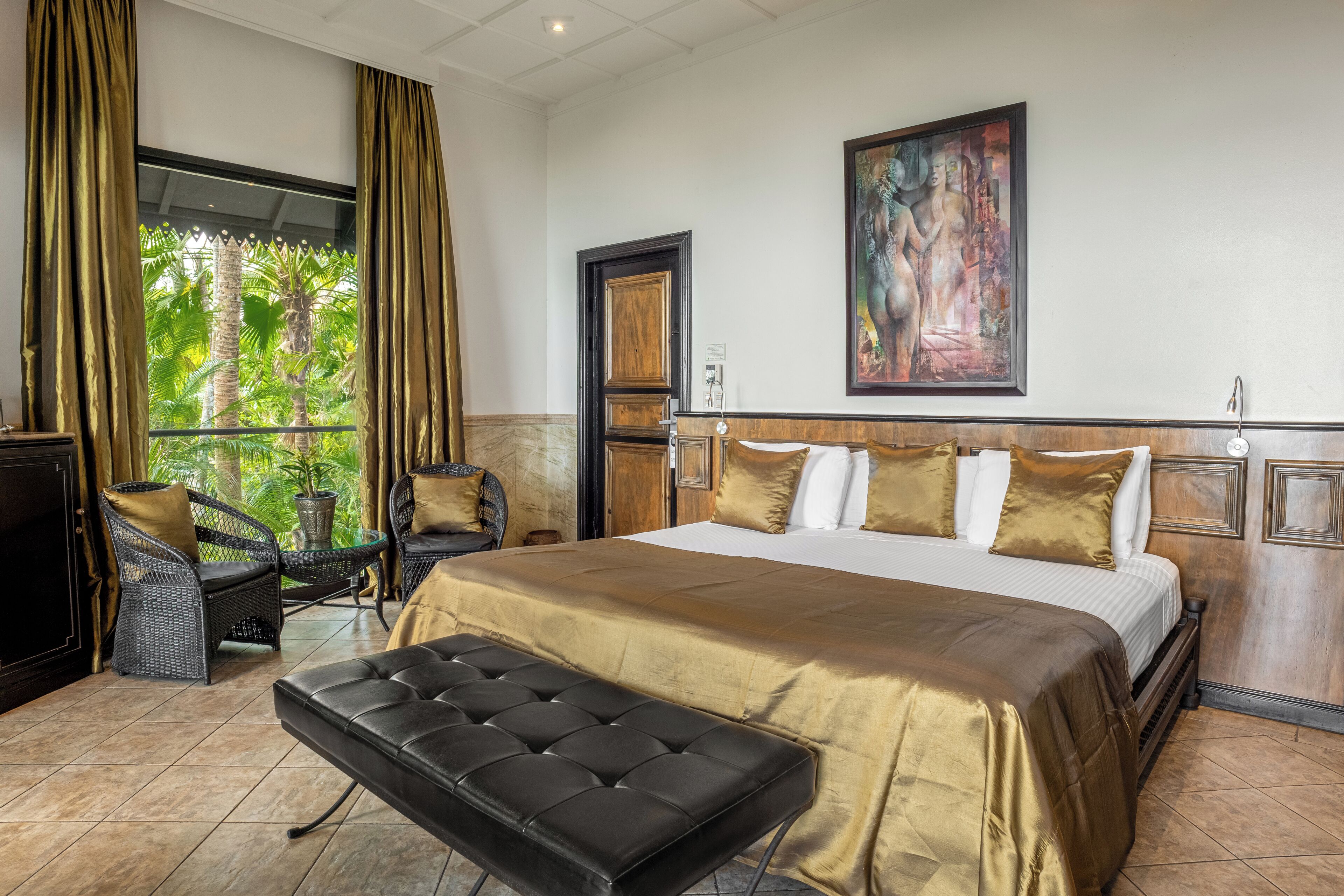 Hotelkamer met een groot bed, een bankje, rieten stoelen, een televisie en een schilderij aan de muur.