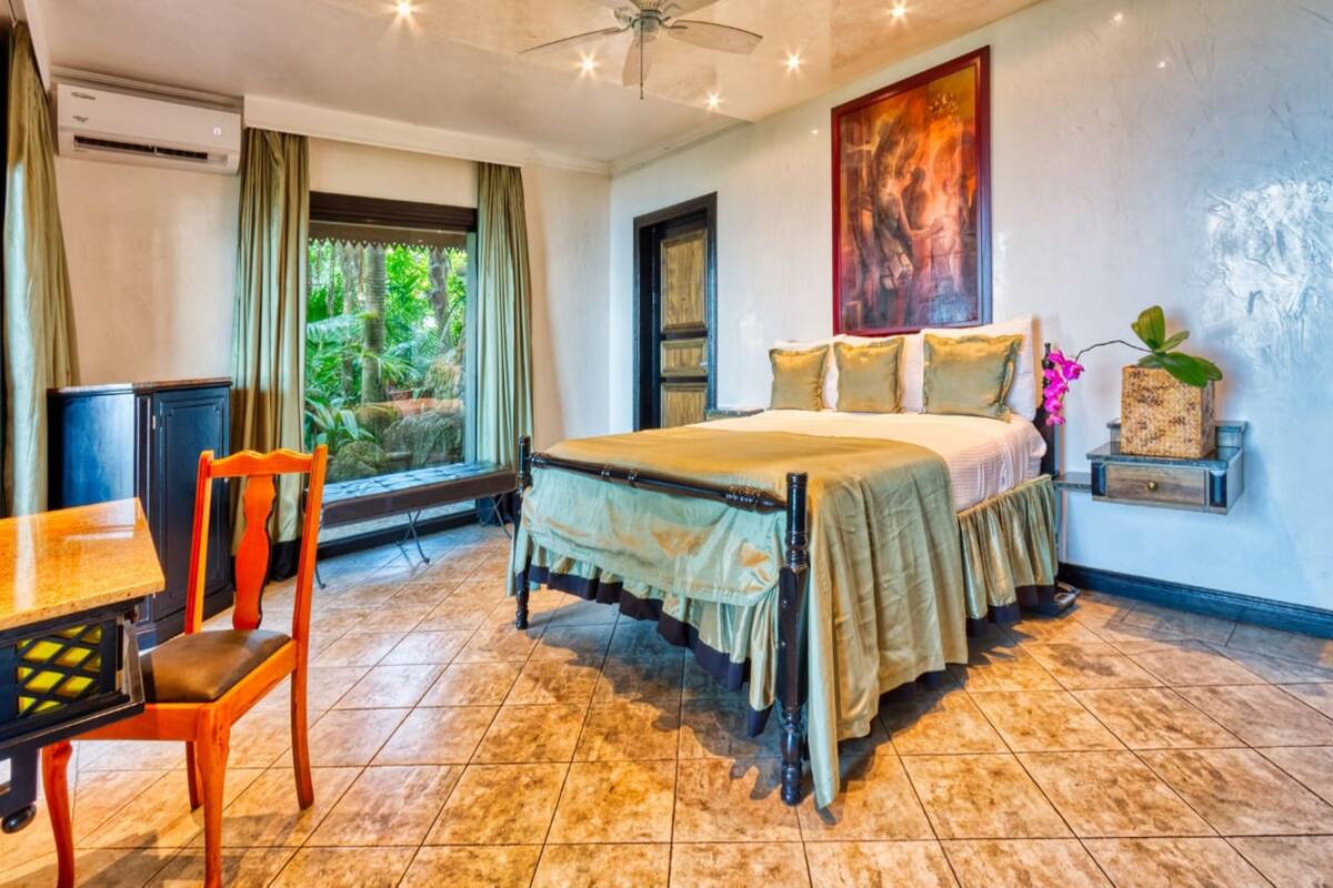 A bedroom with a bed, a desk, and a chair. There is a window with curtains, a ceiling fan, and a painting on the wall.
