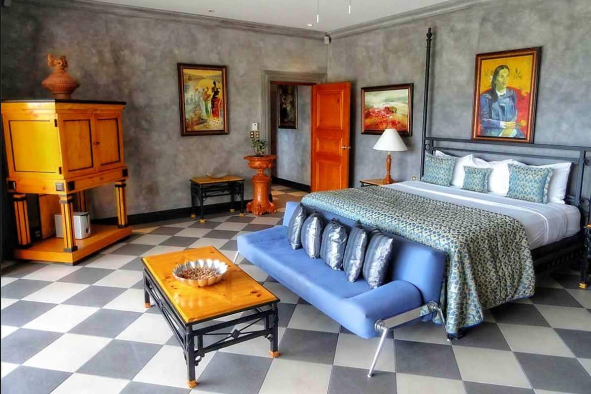 A hotel room with a checkered floor, a bed with patterned bedding, a blue sofa, a wooden cabinet, and artwork on the walls.