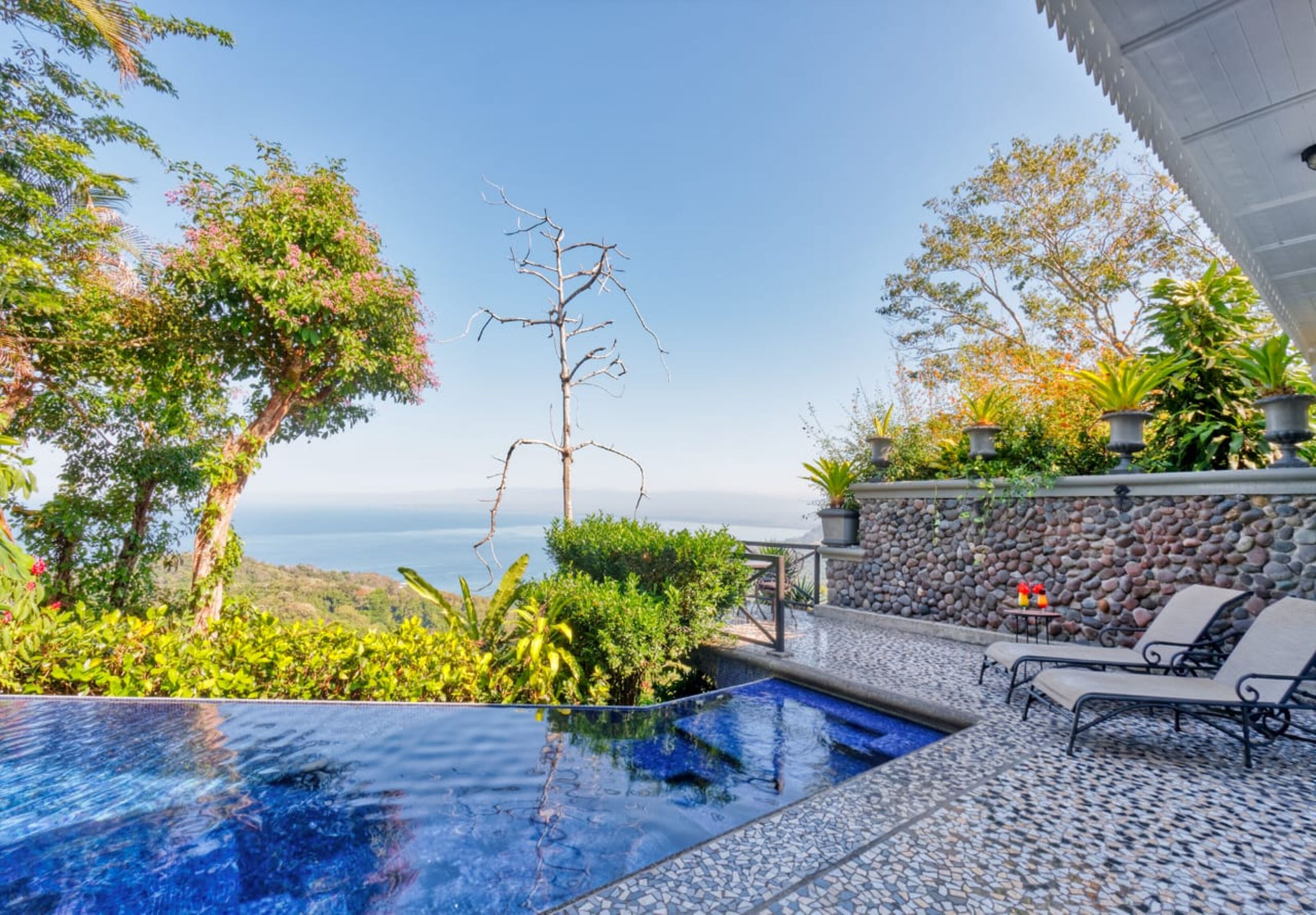 A pool with a clear view of the ocean, surrounded by lush greenery and a stone wall.