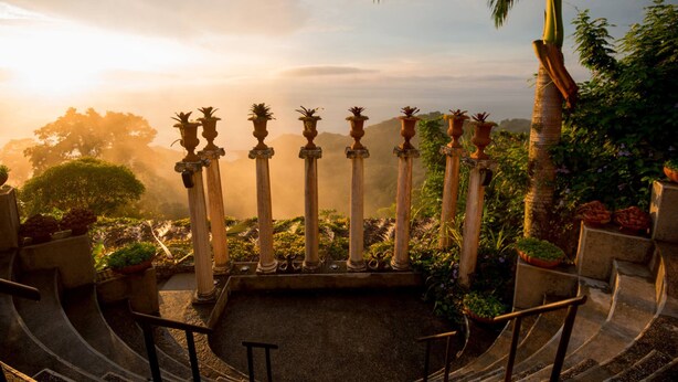 A garden with a central fountain, surrounded by stone columns and potted plants, with a view of distant trees and a clear sky. - Hotel Villa Caletas (Tárcoles)