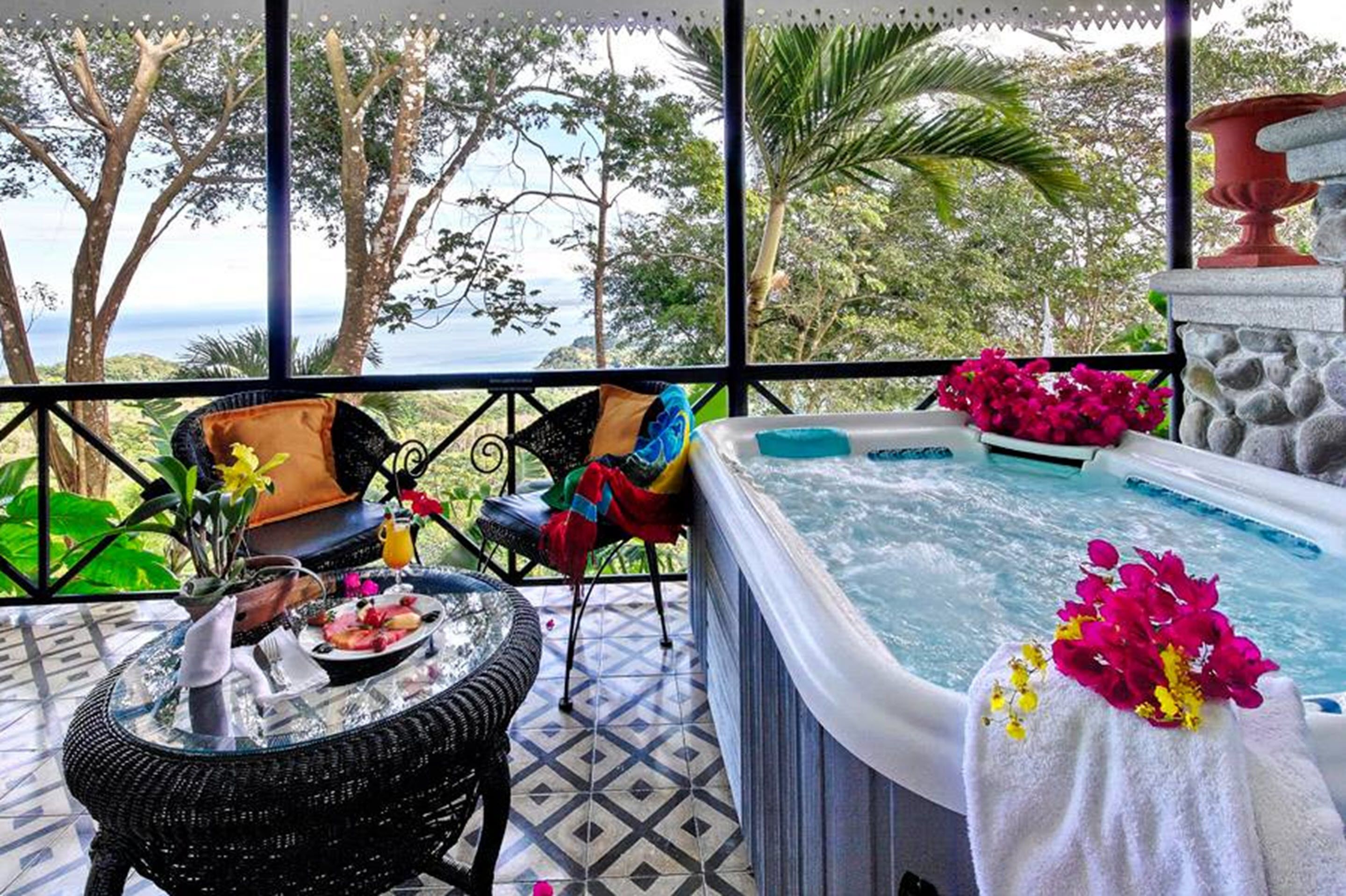 A balcony with a hot tub, wicker chairs, and a dining table with a flower vase and a plate of food.