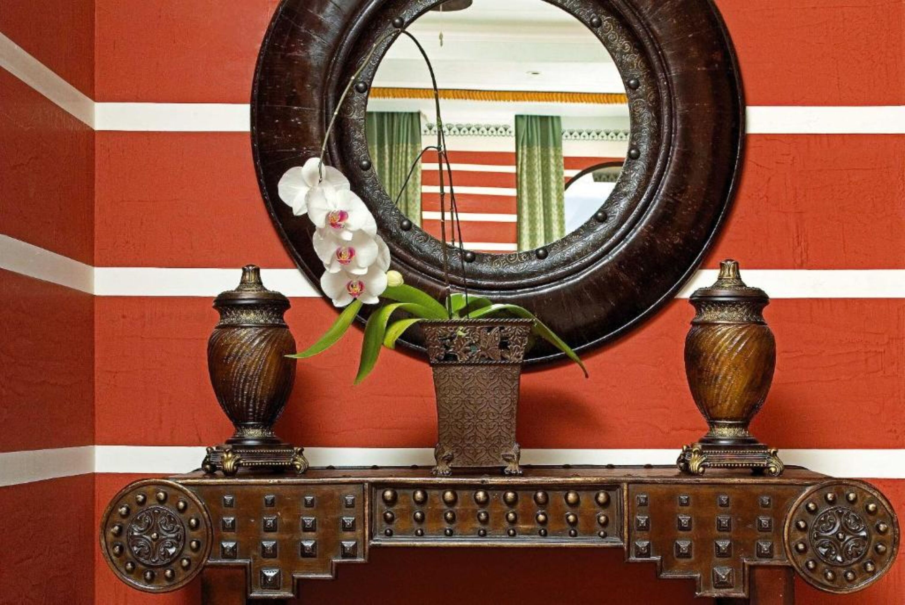 Un miroir décoratif doté d’un cadre rond, encadré de deux vases richement décorés et agrémenté d’une petite plante en pot aux fleurs blanches.