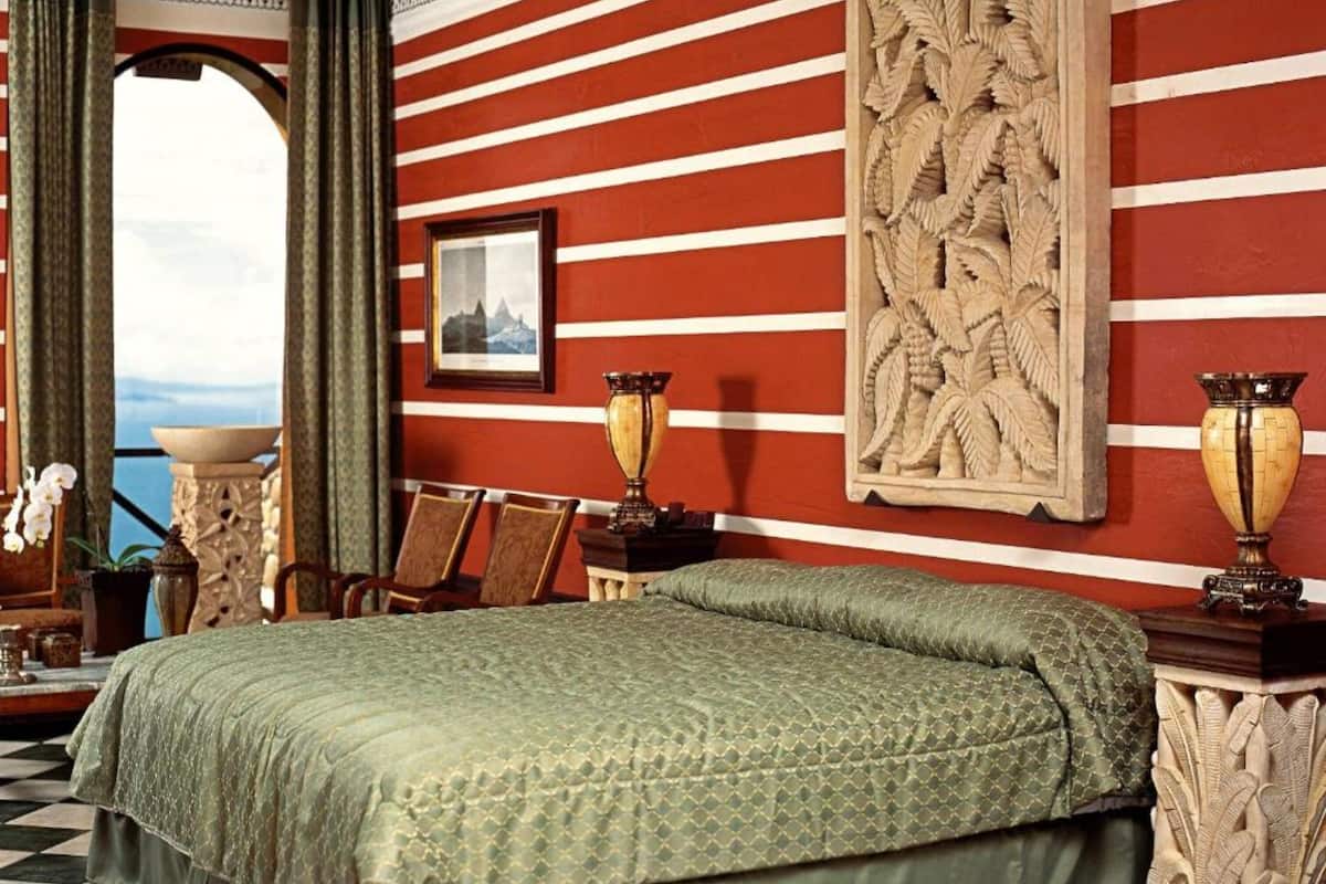 A room with a large bed, a view through an arched window, a framed picture on the wall, and a decorative panel.