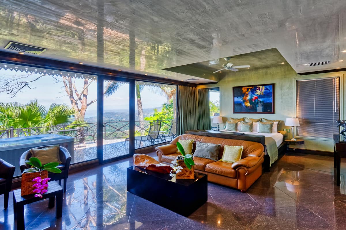 A spacious room with a large bed, a sofa, and a coffee table. There is a balcony with a view of the outdoors.