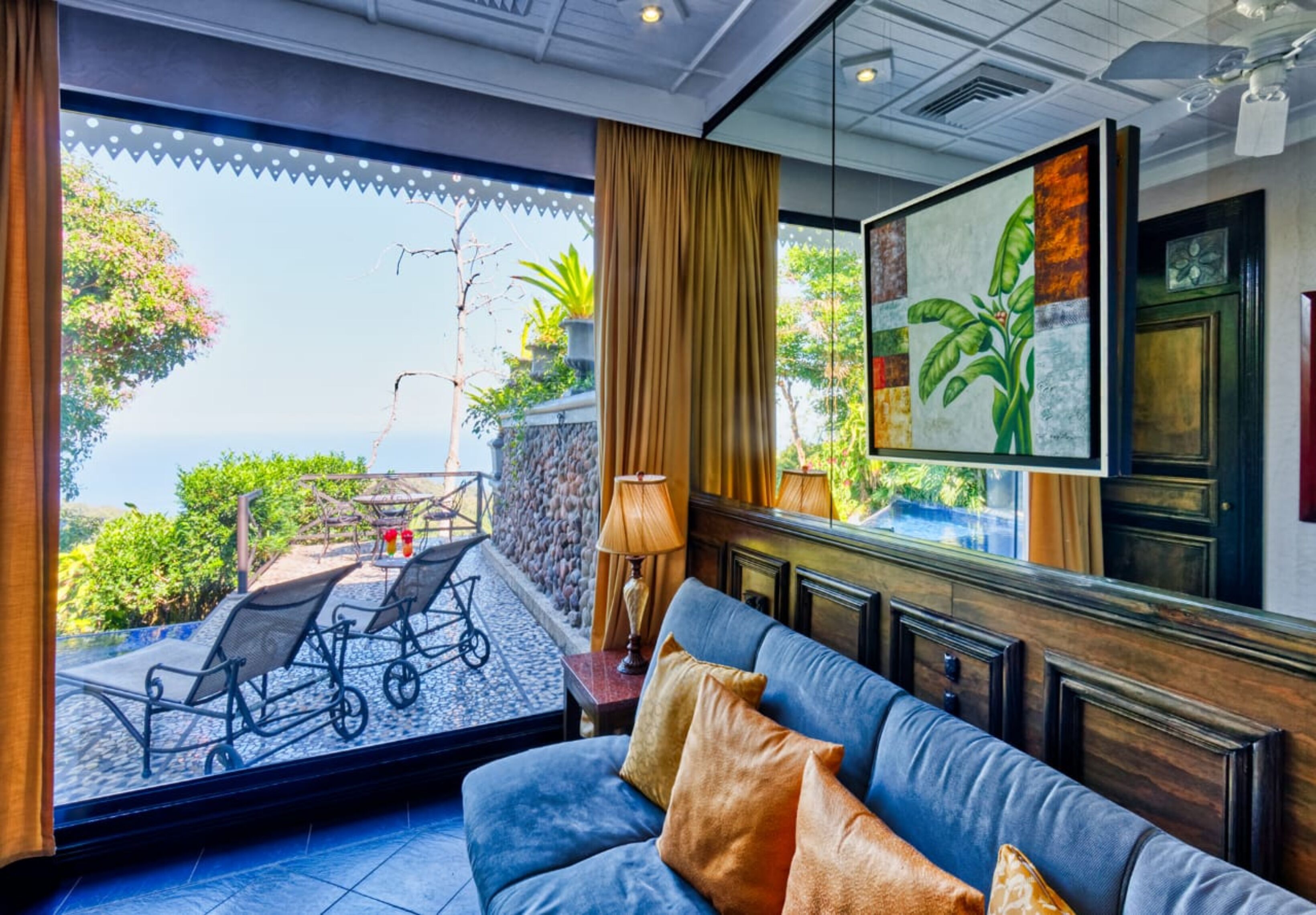 A room with a blue sofa, a view of the outdoors, a painting on the wall, and a window with curtains.