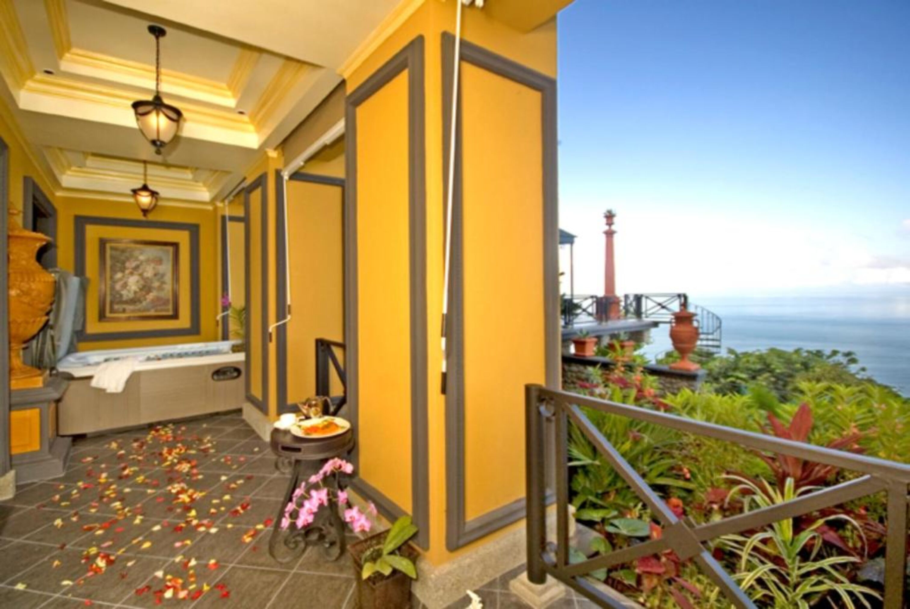 A hotel corridor with a view of the sea, a bench, a table with flowers, and a railing with plants.