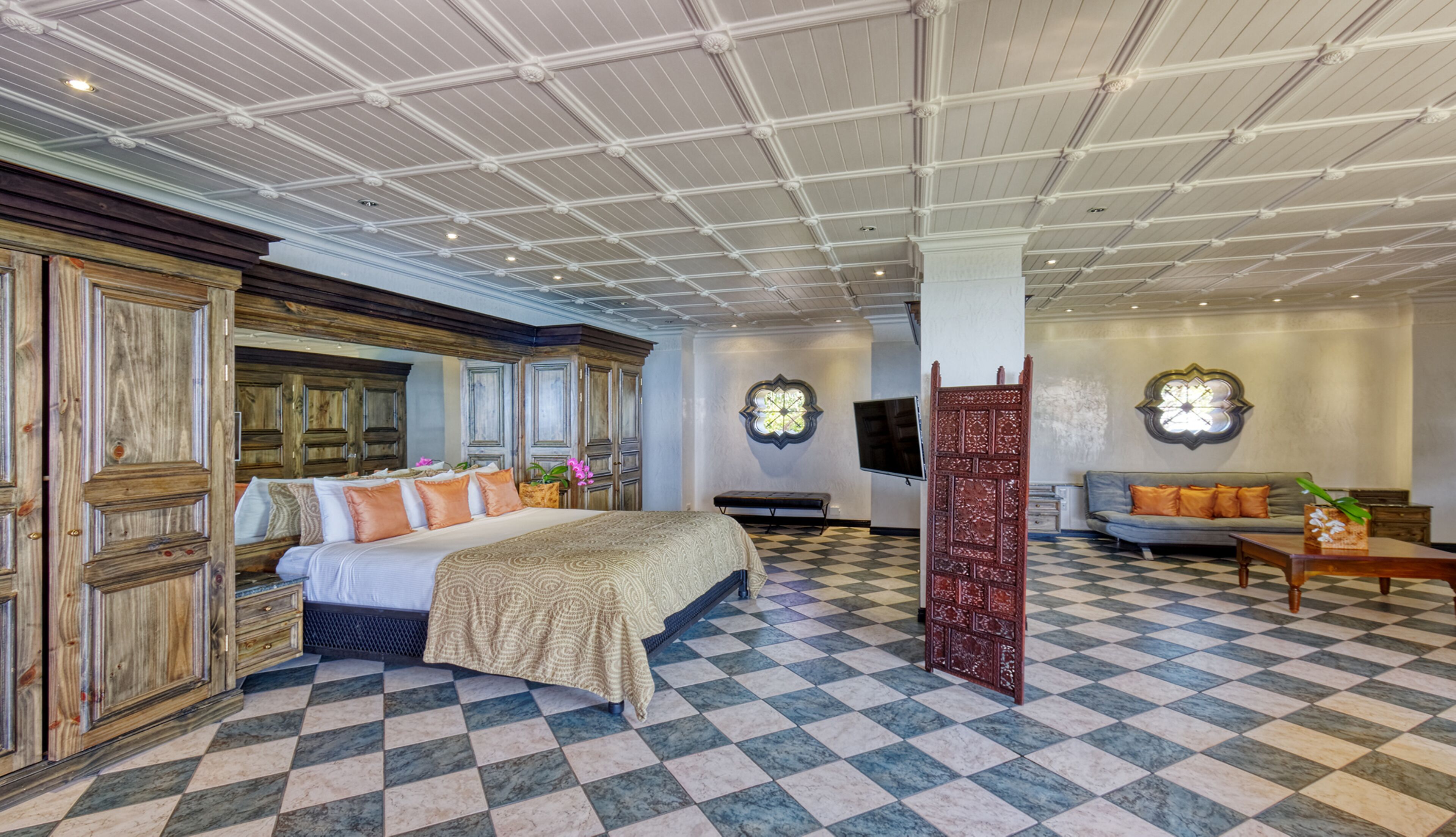 A spacious room with a large bed, a sofa, and a TV. The room has a checkered floor and a high ceiling with decorative molding.