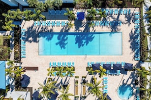 An aerial view of a resort with a large rectangular pool surrounded by rows of blue lounge chairs and umbrellas, with palm trees and a clear sky.