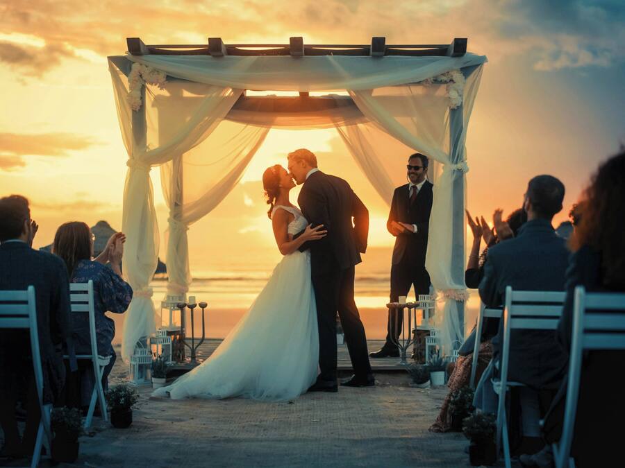 Una cerimonia nuziale con invitati seduti su sedie bianche, una coppia che si bacia sotto una tettoia e un tramonto sullo sfondo.