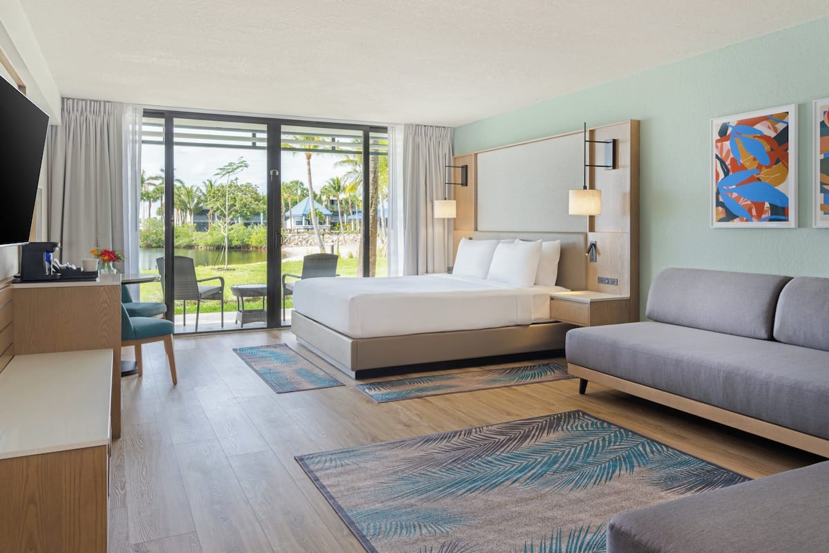 A hotel room with a bed, a sofa, a desk, and a TV. There is a balcony with a view of palm trees and a pool.