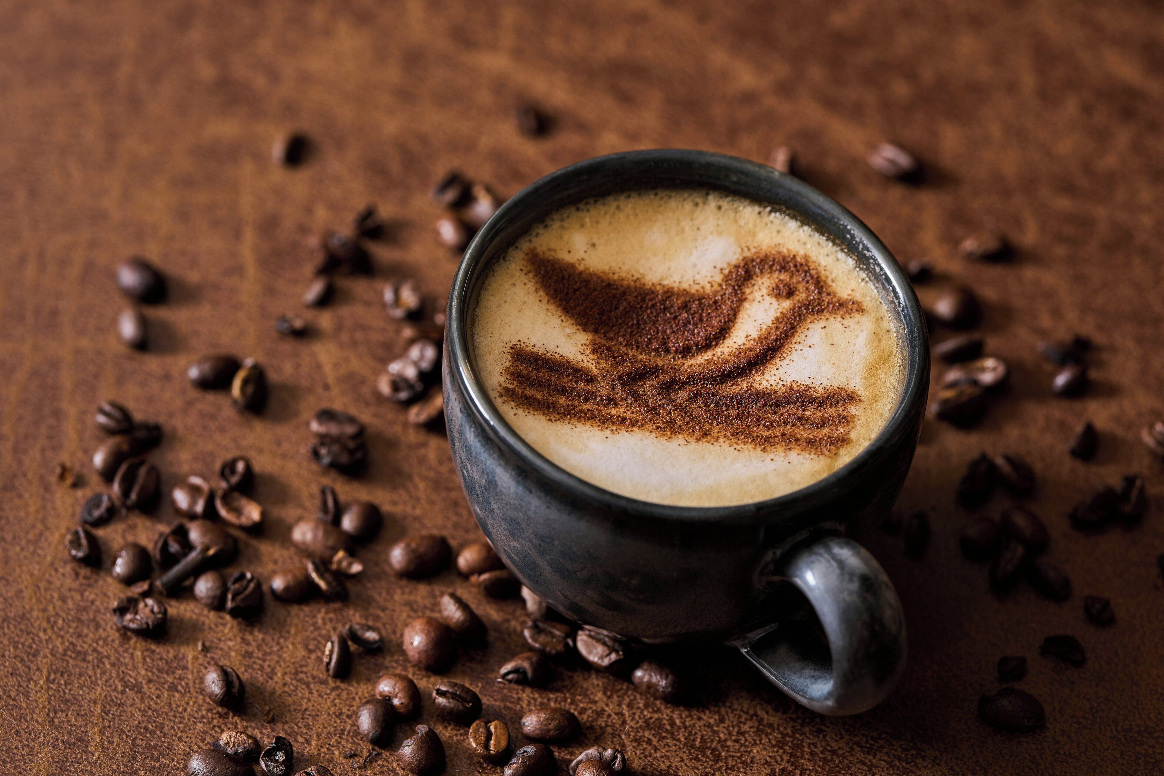 Una taza de café con un dibujo de ave en espuma, sobre una superficie texturizada rodeada de granos de café.