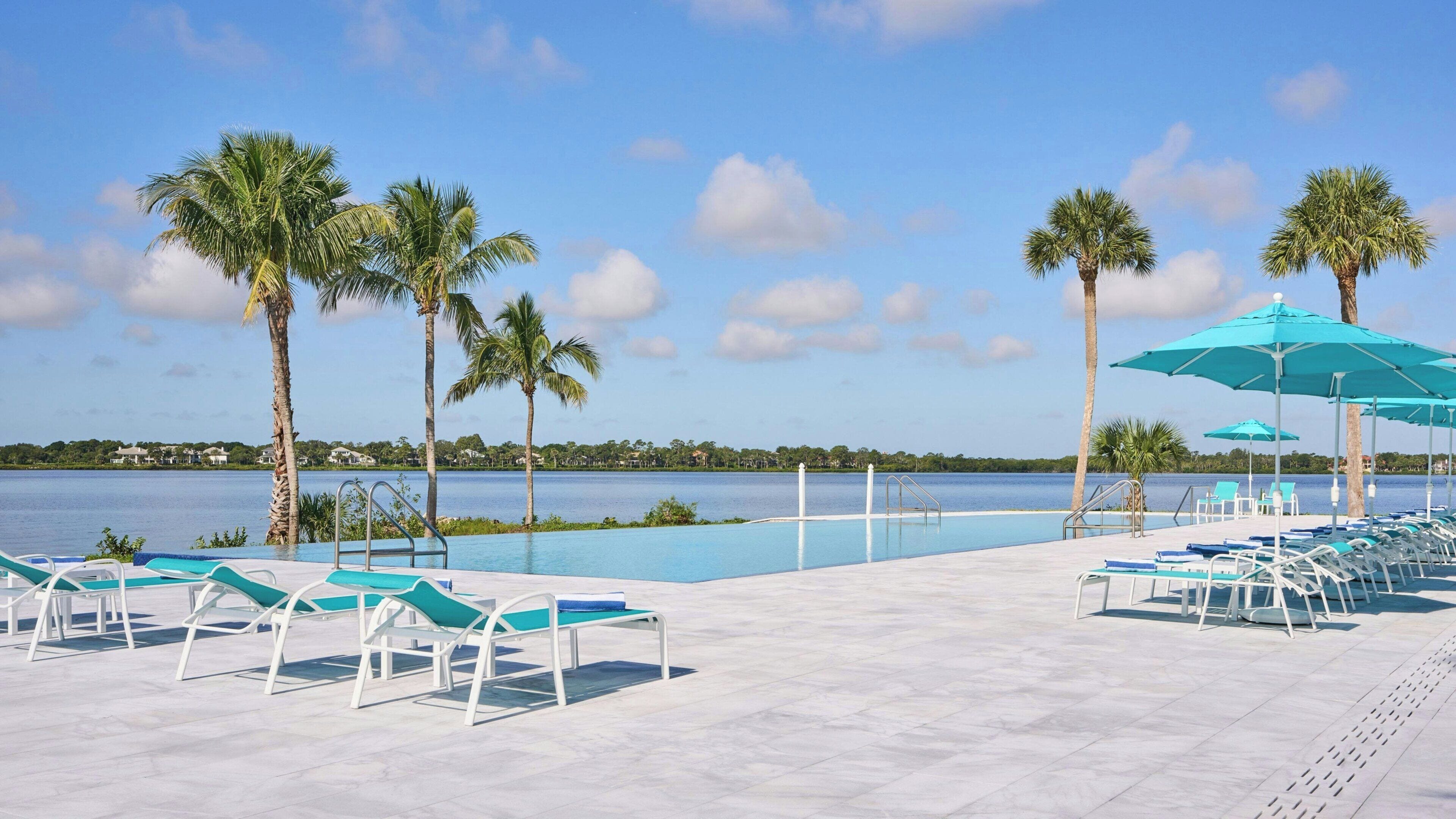 A poolside area with lounge chairs and umbrellas, palm trees, and a view of a lake.