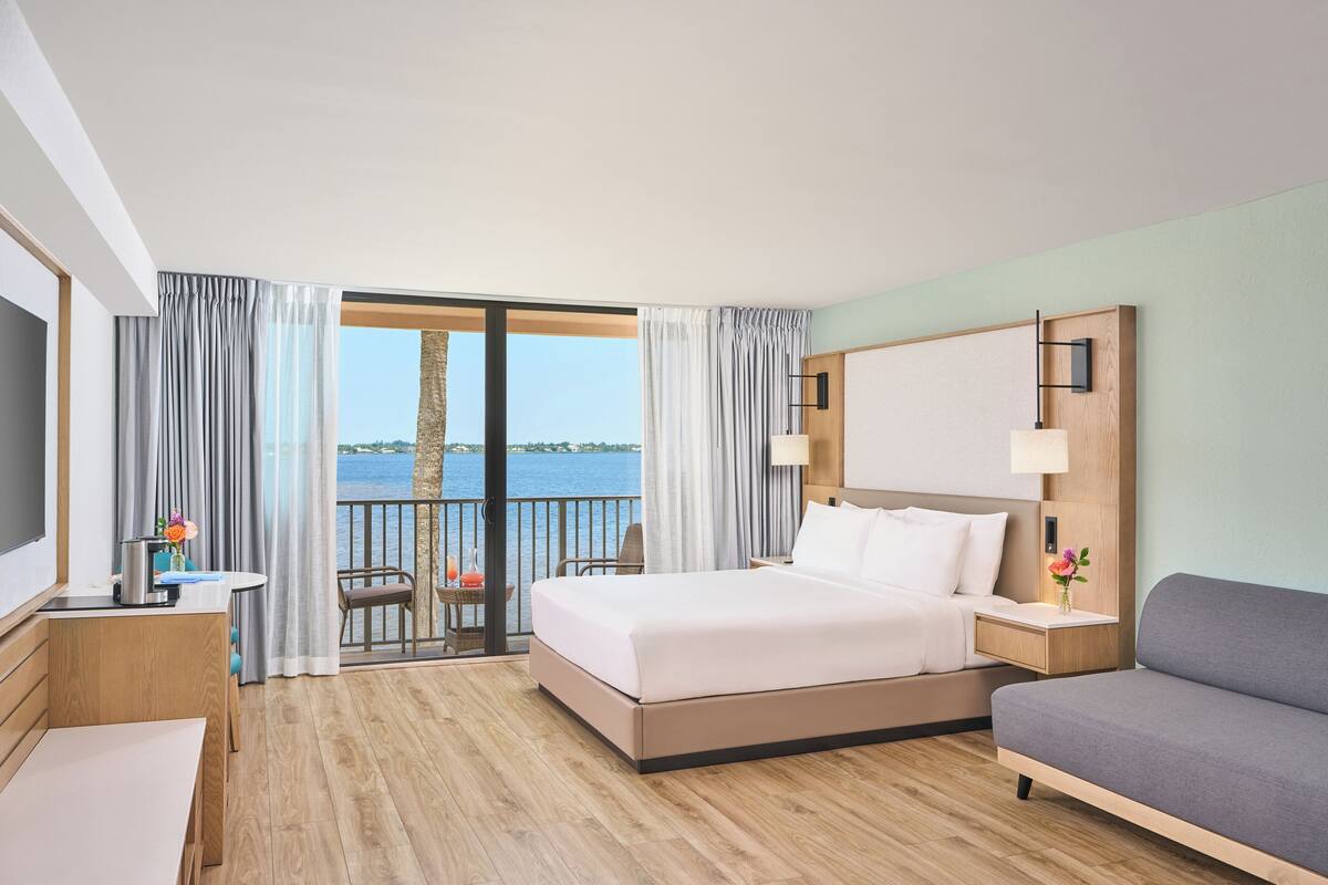 A hotel room with a bed, a sofa, a desk, and a TV. There is a balcony with a view of the water.
