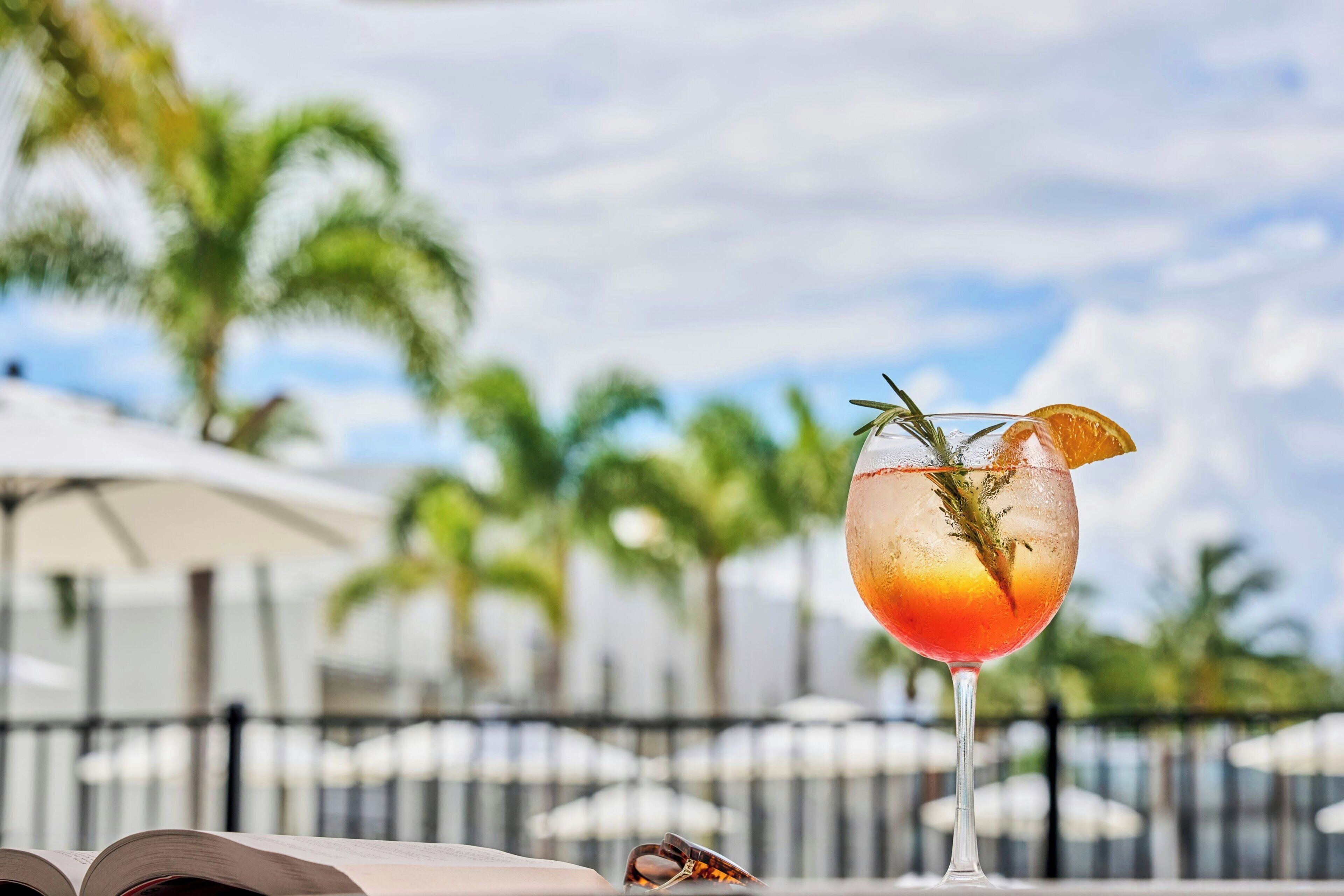 A cocktail glass with a garnish on a table with a book, palm trees, and umbrellas in the background.