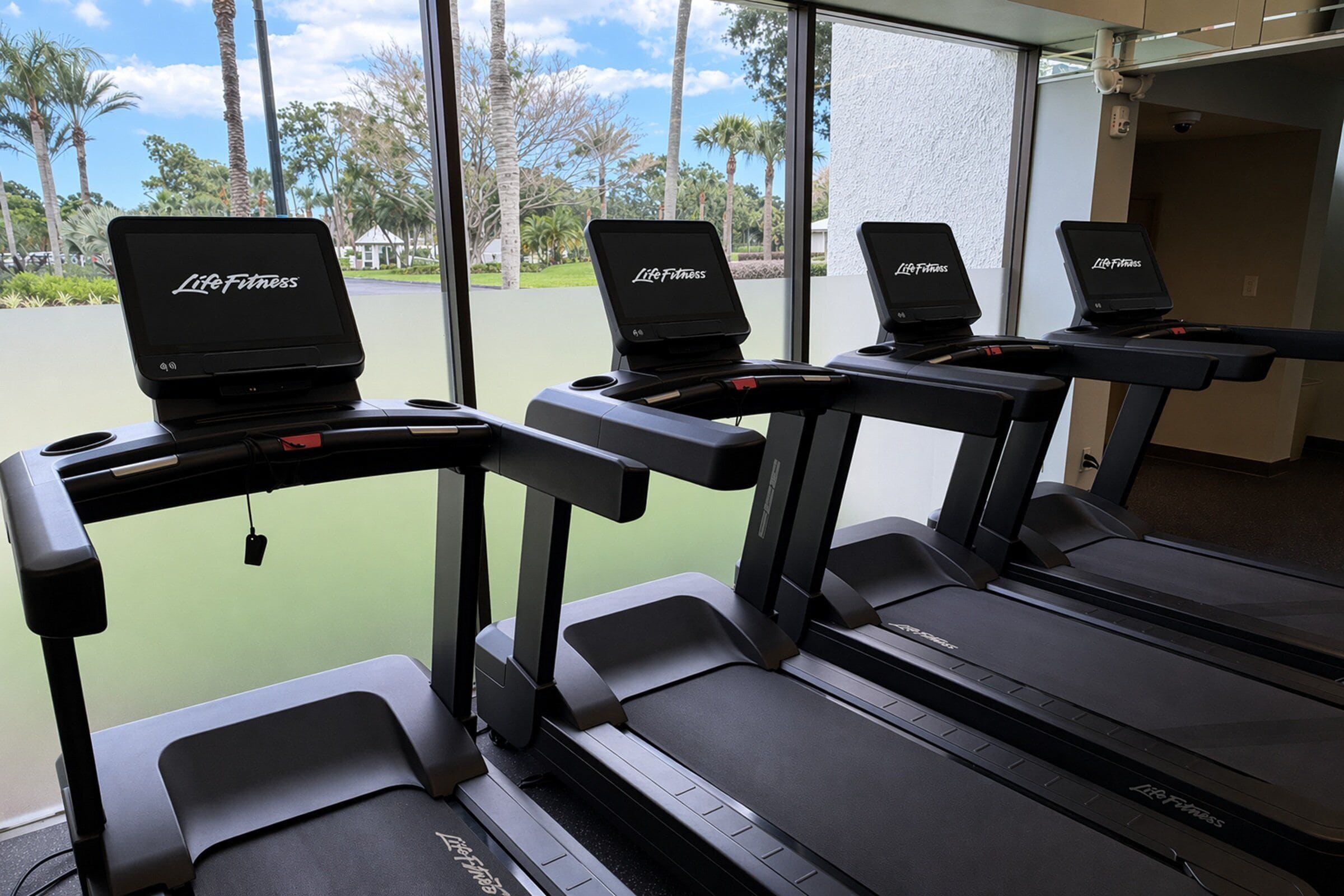 Drie loopbanden met 'Life Fitness'-logo, geplaatst in een ruimte met grote ramen met uitzicht op een groen buitengebied met palmbomen.