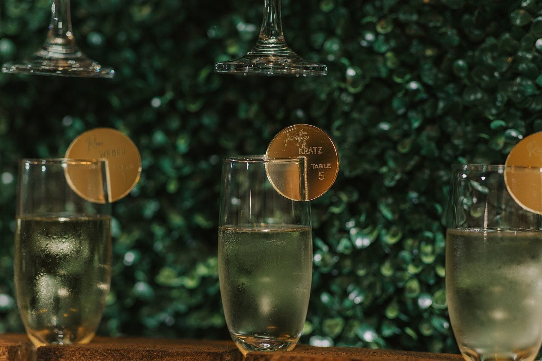 Three glasses of a clear liquid, possibly a cocktail, with a gold-colored coaster on each. The background is a blurred green foliage.