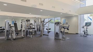 Un gimnasio con diversas máquinas de ejercicio, un reloj de pared y una ventana con vistas a palmeras.