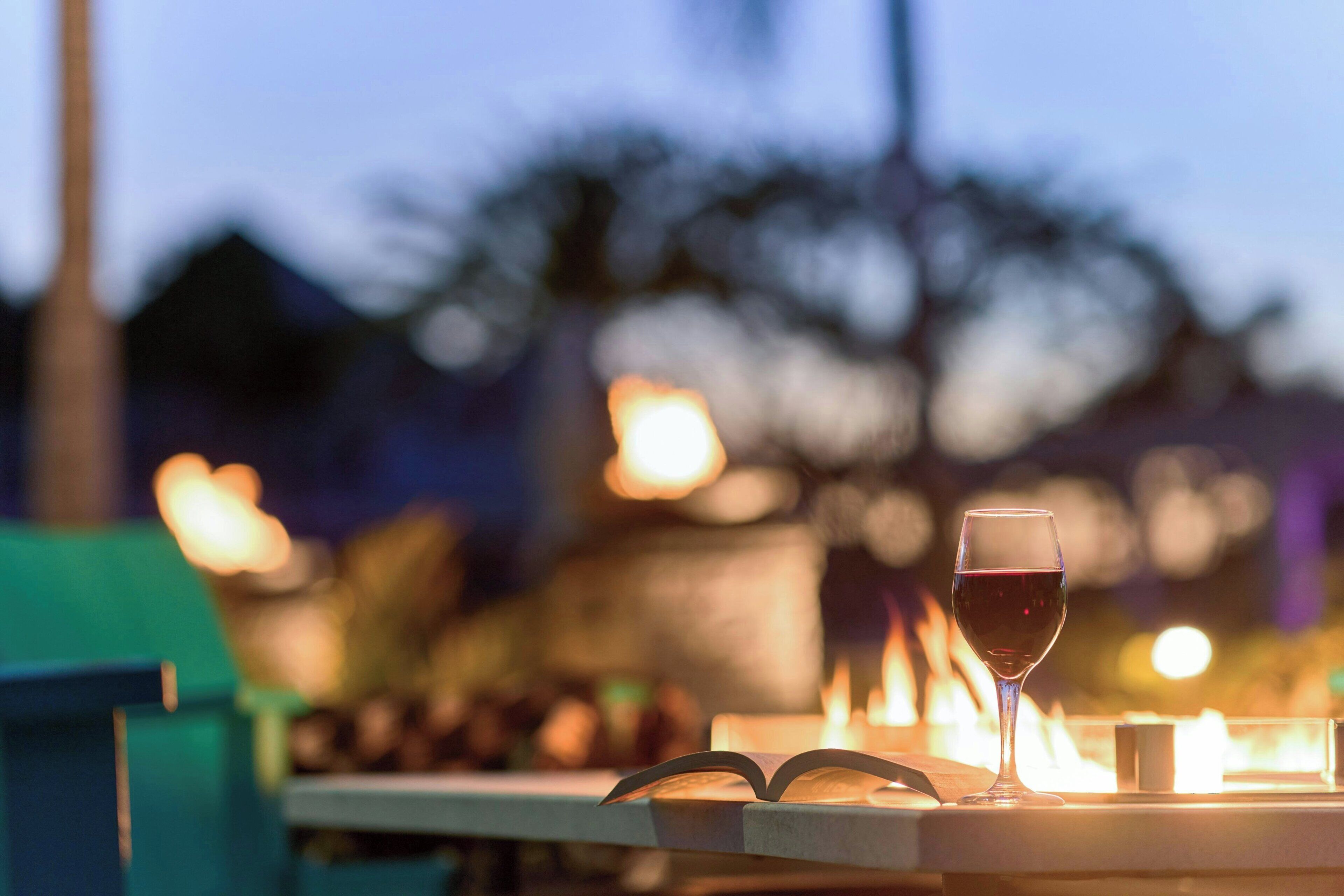 A table with a glass of red wine and a book, with a blurred background of lights and trees.