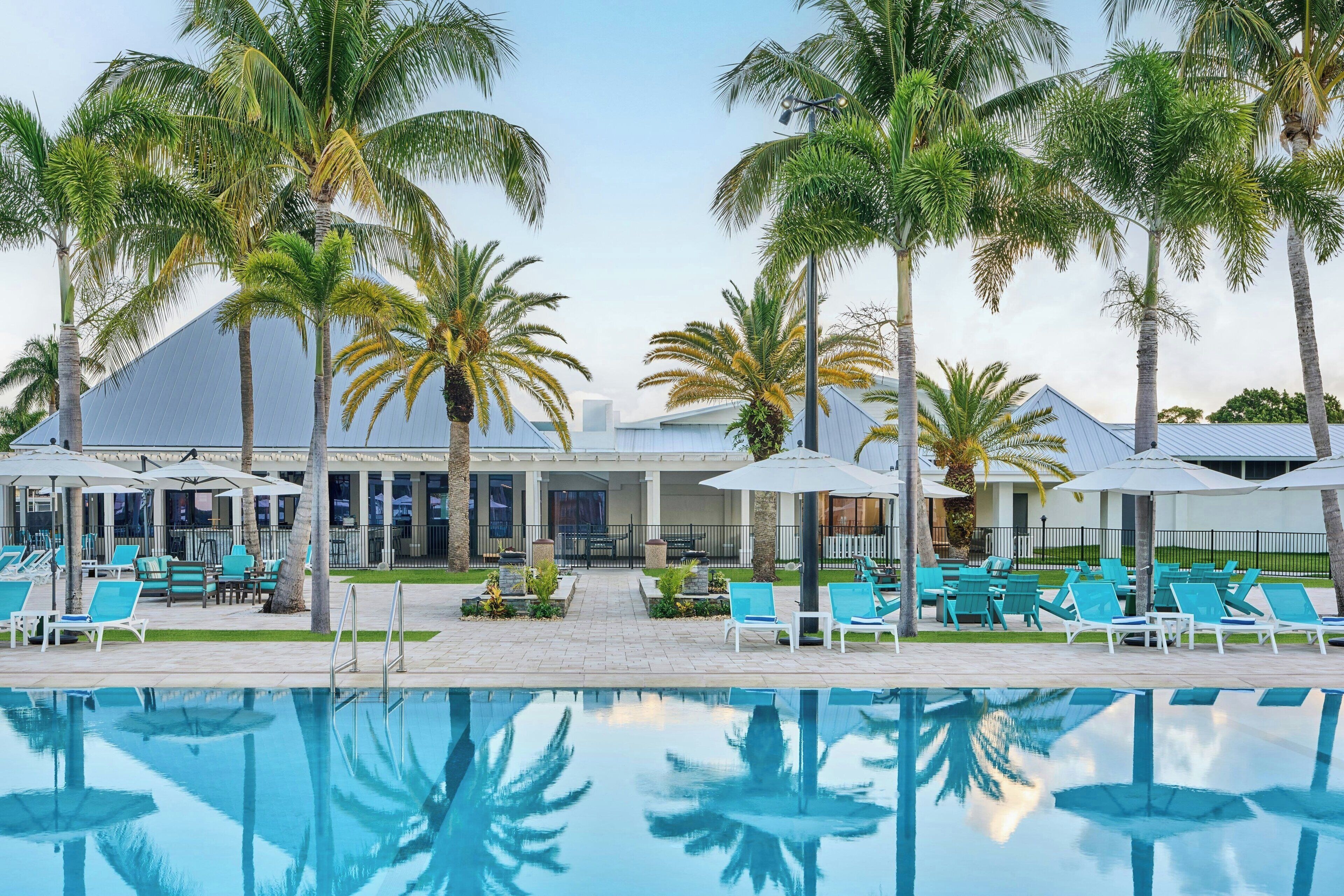 A poolside area with lounge chairs and umbrellas, palm trees, and a building with a blue roof.