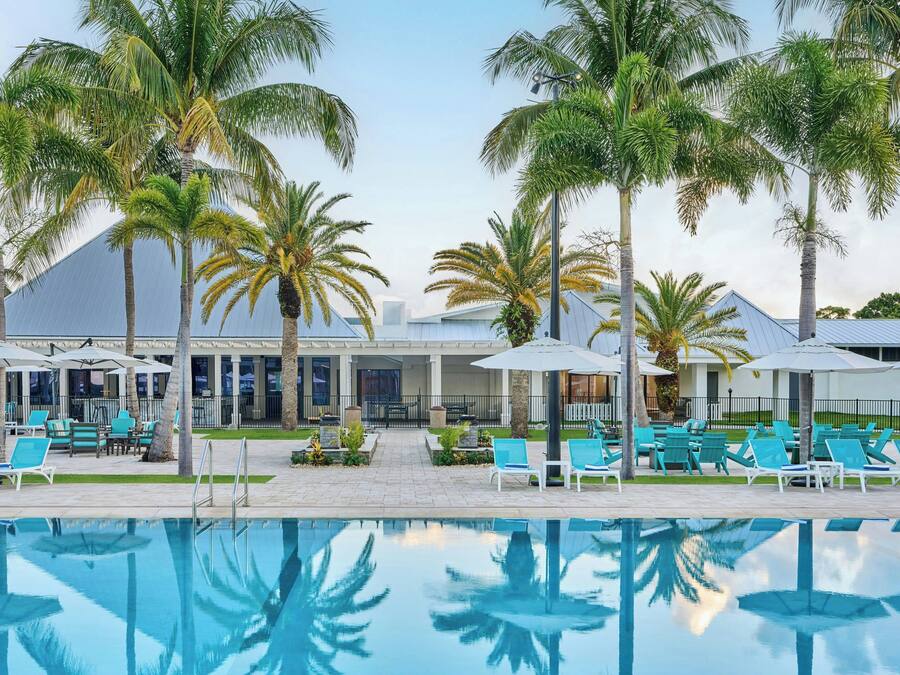 A poolside area with lounge chairs and umbrellas, palm trees, and a building with a blue roof.