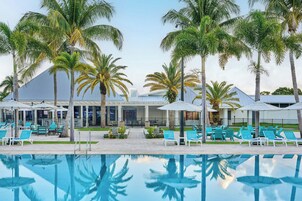 Zona junto a la piscina con sillones de descanso y sombrillas, palmeras y un edificio con techo azul.