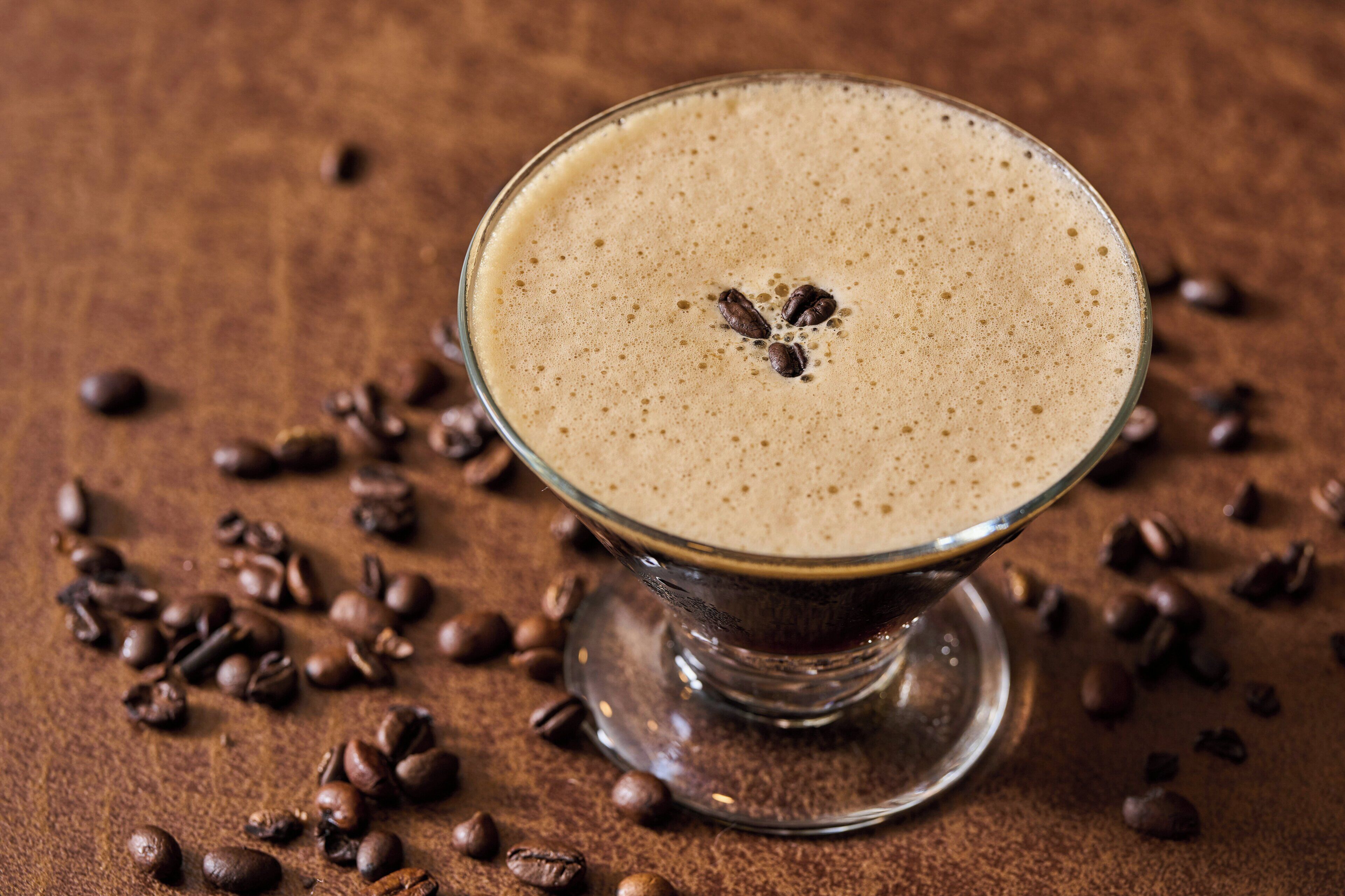 A glass of coffee with a frothy top, surrounded by coffee beans on a textured surface.