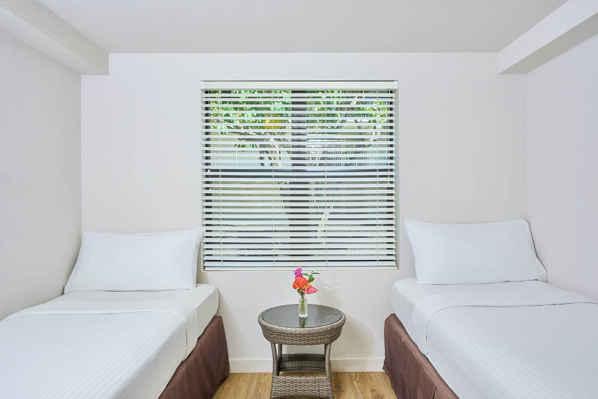 A room with two single beds, a small round table with a vase, and a window with blinds.