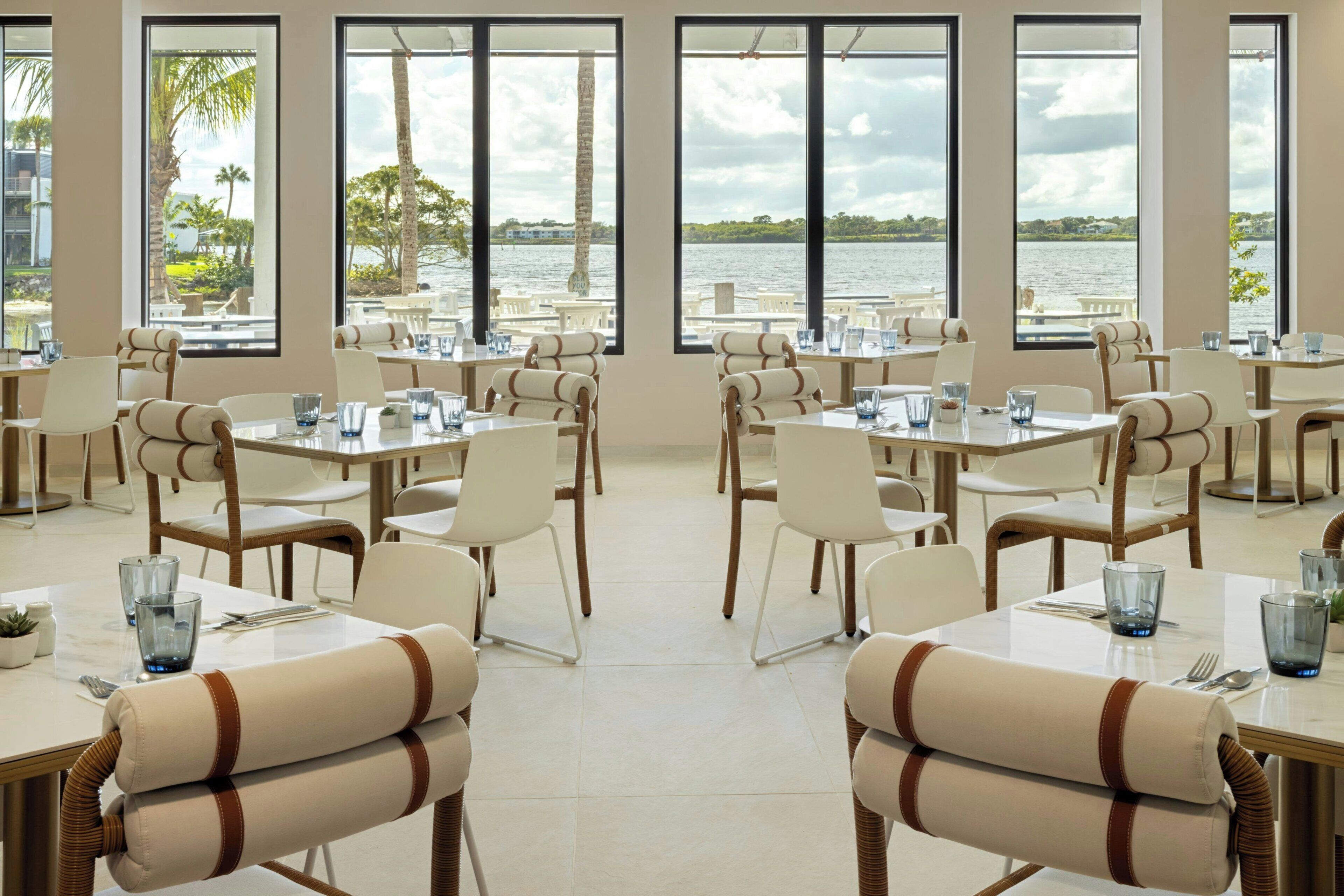 Un restaurante con varias mesas y sillas, amplias ventanas que ofrecen vista a un cuerpo de agua y follaje.