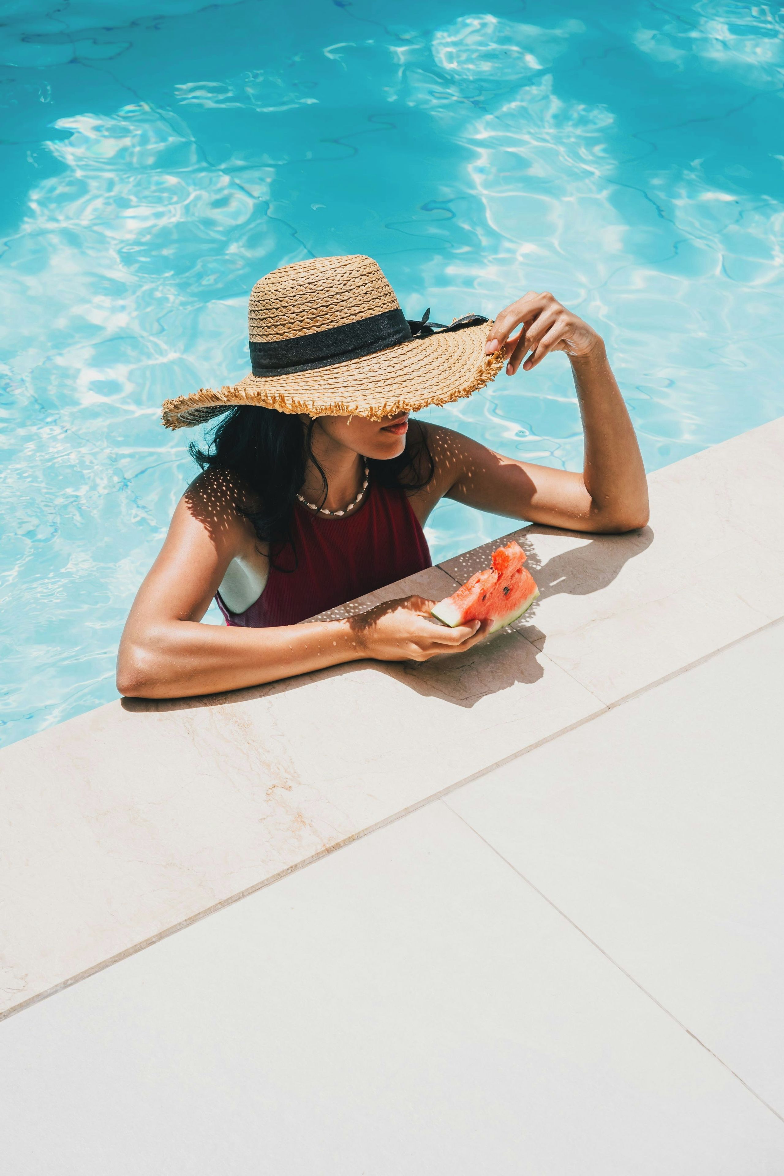 Une personne assise au bord d’une piscine, portant un chapeau et tenant une tranche de pastèque.