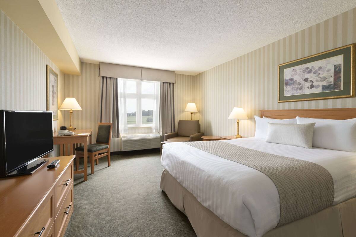 Ein Hotelzimmer mit einem großen Bett, einem Schreibtisch mit Fernseher, einem Sessel und einem Fenster mit Vorhängen.