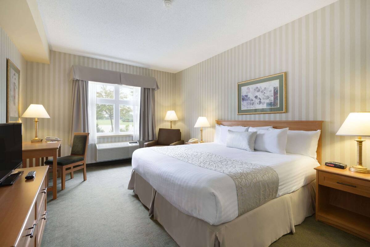 Ein Hotelzimmer mit einem großen Bett, zwei Nachttischen mit Lampen, einem Schreibtisch mit Stuhl, einem Fernseher und einem gerahmten Bild an der Wand.