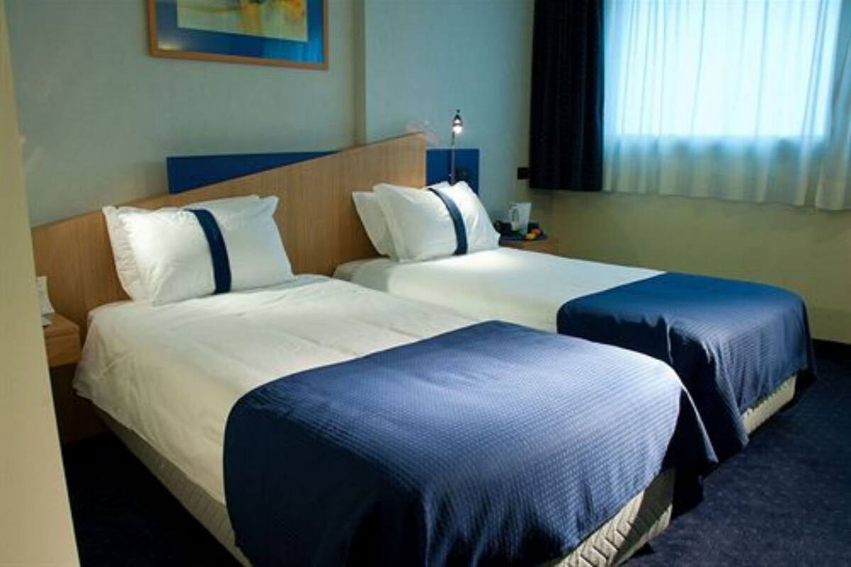 A hotel room with two beds, each with a blue bedspread and white pillows. There is a nightstand with a lamp and a small table with a cup and fruit.