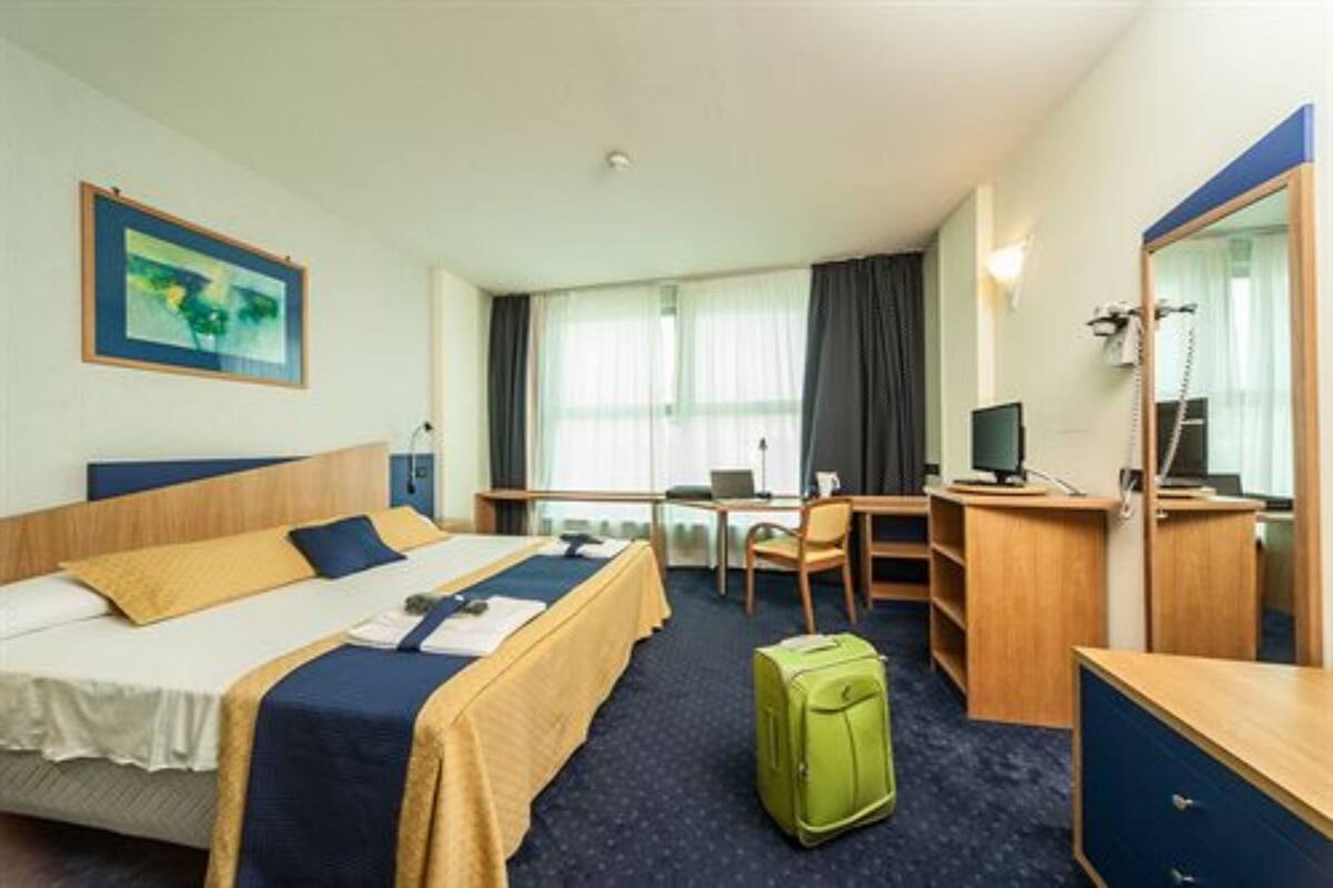 A hotel room with a bed, desk, chair, and a green suitcase.
