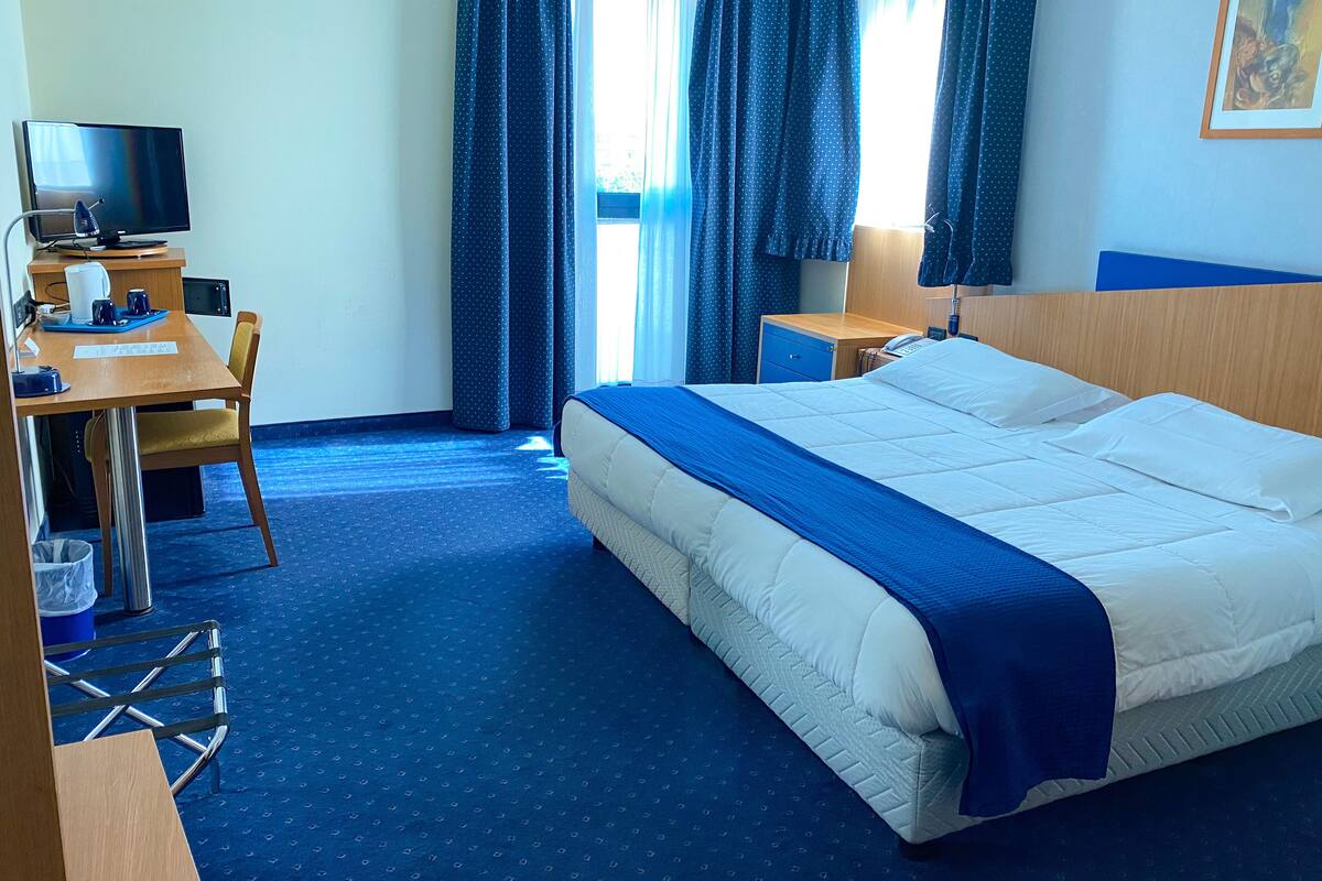 A hotel room with a bed, a desk with a computer, a chair, a small table, and a window with curtains.