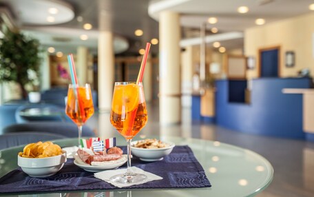 A table with a glass of orange juice, a glass of red wine, a bowl of chips, and a plate of meatballs, set on a glass table in a hotel lobby.